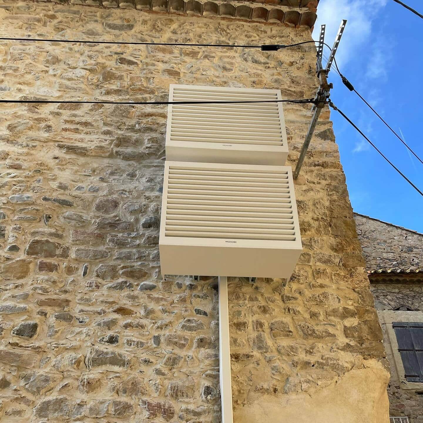Deux caches d'unité extérieure de climatisation DECO'Clim beige, à persiennes, fixés sur un mur en pierres apparentes sous un ciel bleu.