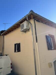 Façade d'une maison claire sous ciel bleu, avec unité extérieure Mitsubishi Electric, gouttière, antenne TV et fourgonnette Peugeot Partner.