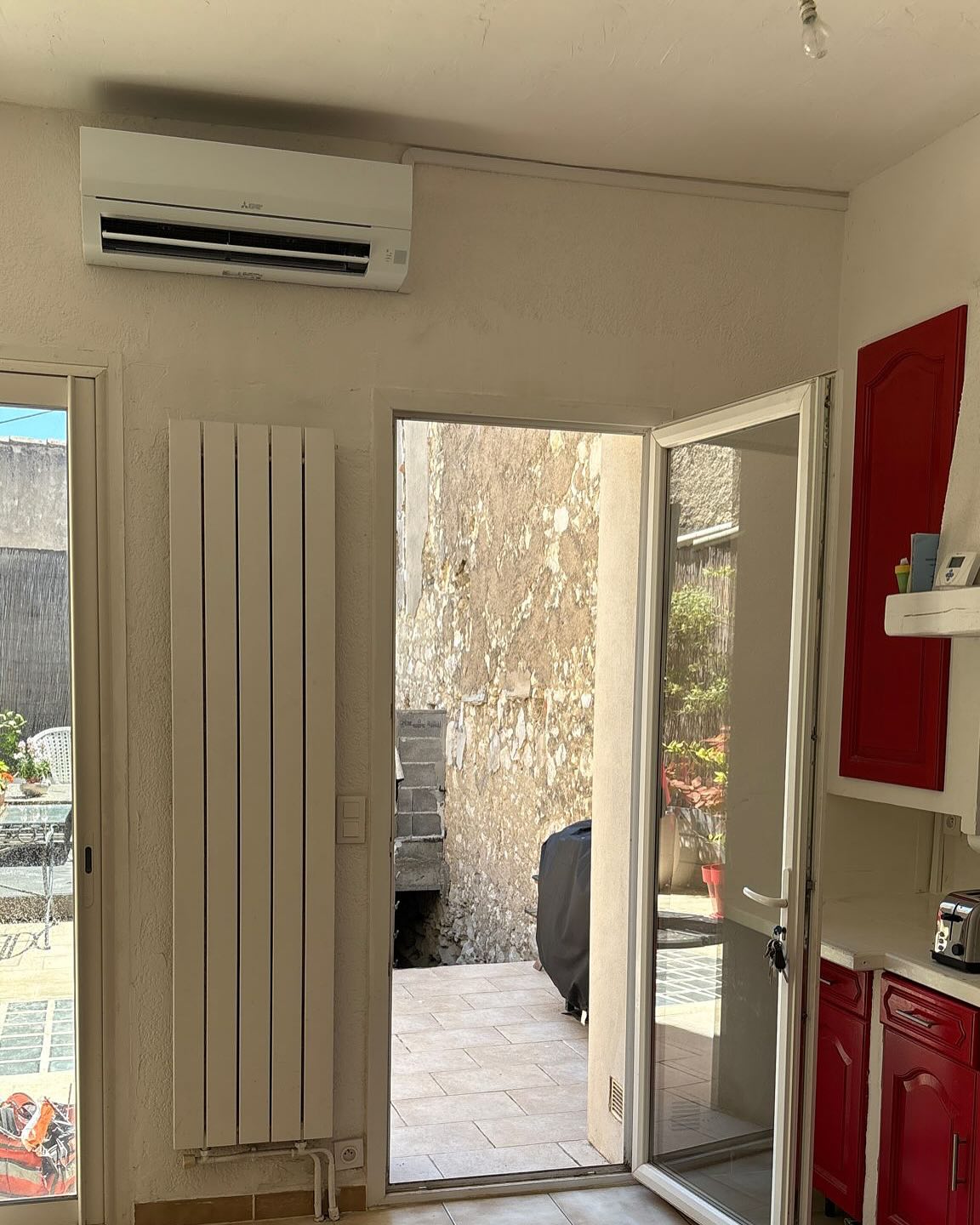 Intérieur avec climatiseur Mitsubishi, radiateur blanc vertical, porte ouverte sur terrasse dallée, et cuisine avec placards rouges.