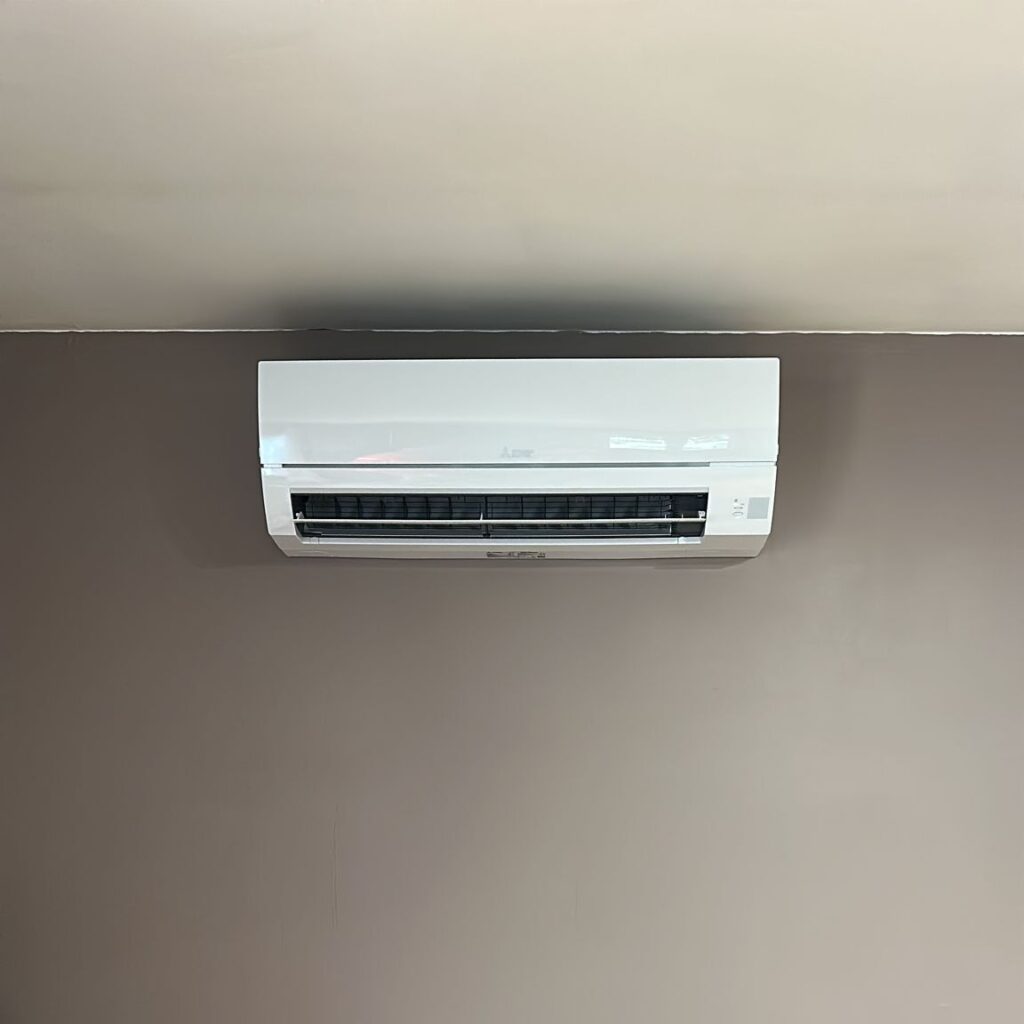 Climatiseur mural Mitsubishi blanc installé sous un plafond clair, soufflant de l'air sur un mur gris-taupe.