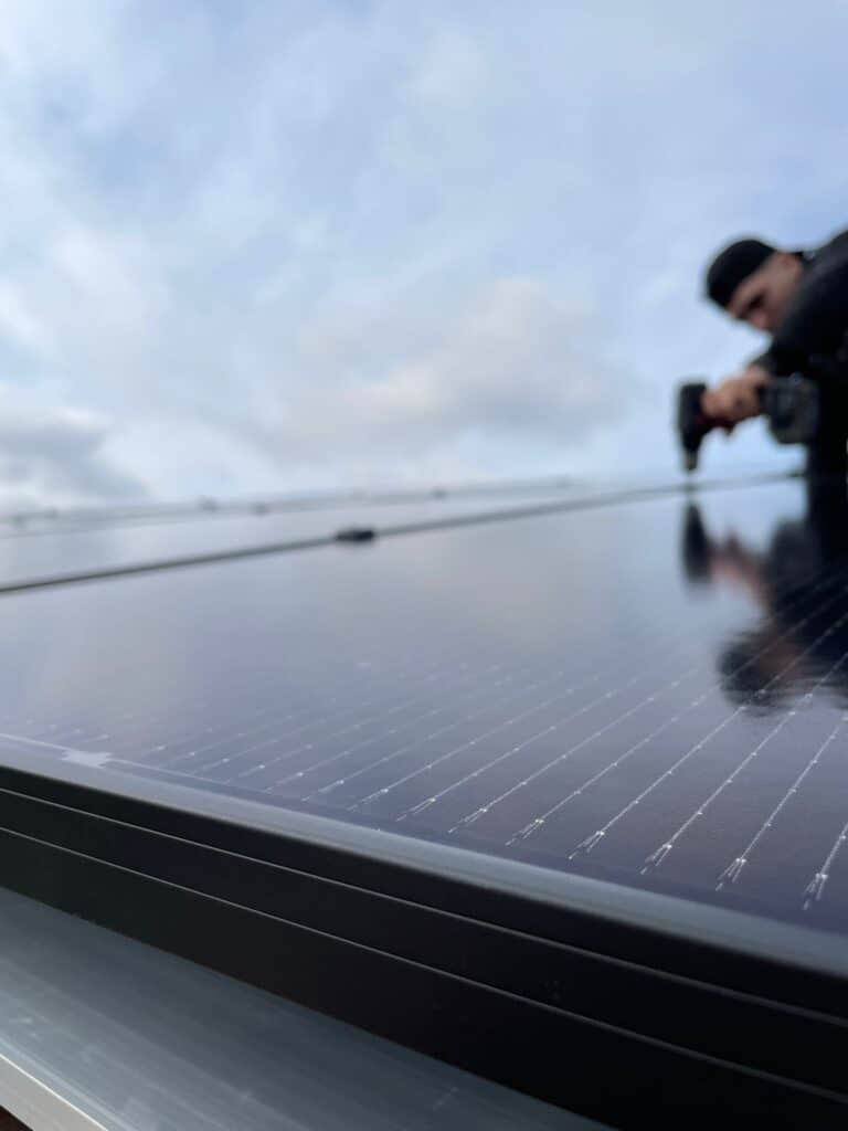 Installation solaire : Pose de panneaux photovoltaïques Installateur de panneaux solaires, casquette noire, travaille sur des modules brillants en gros plan, ciel nuageux en arrière-plan.