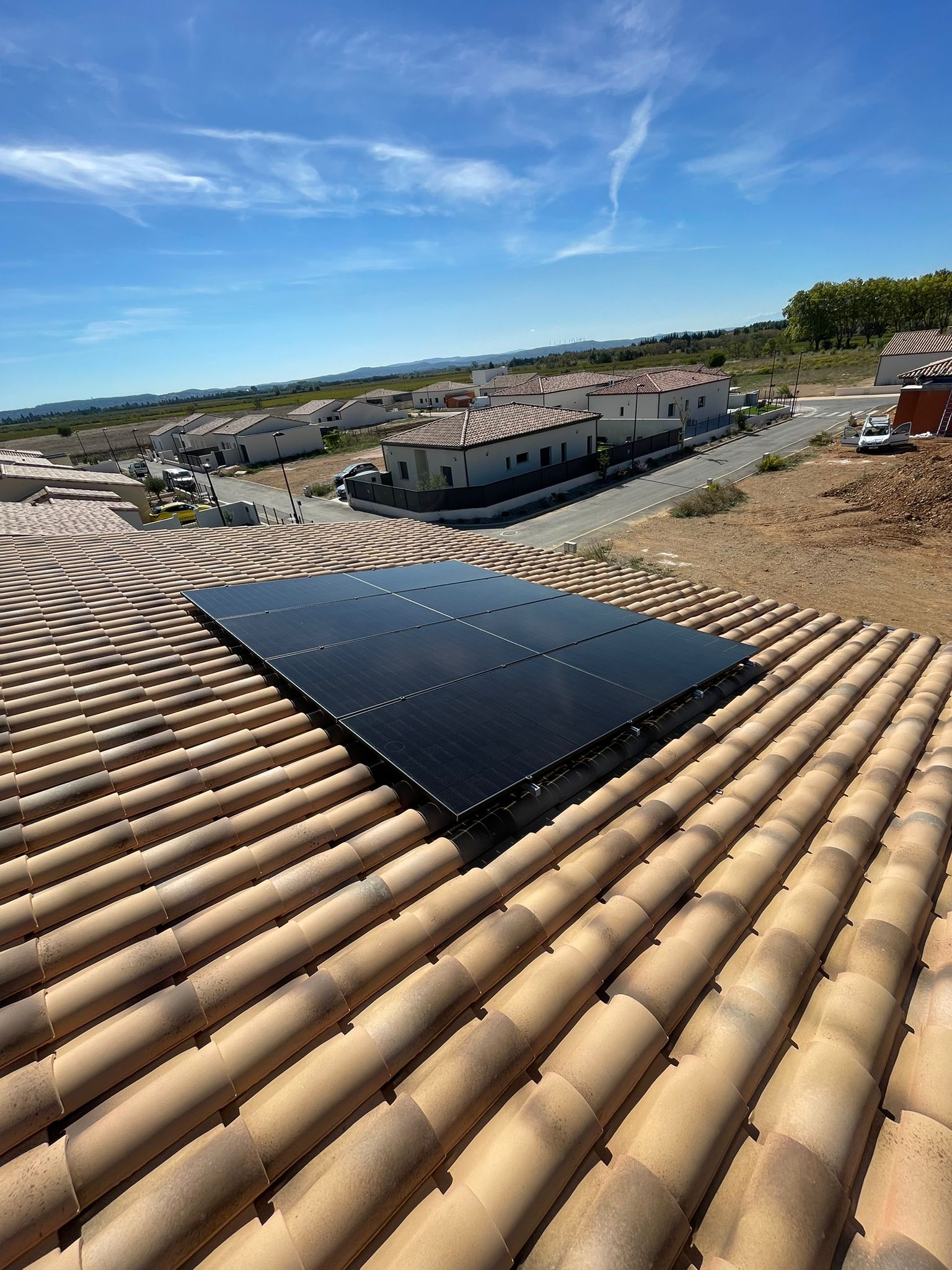 Panneaux solaires noirs installés sur un toit en tuiles romanes couleur terre cuite, surplombant un lotissement récent sous un ciel bleu clair.