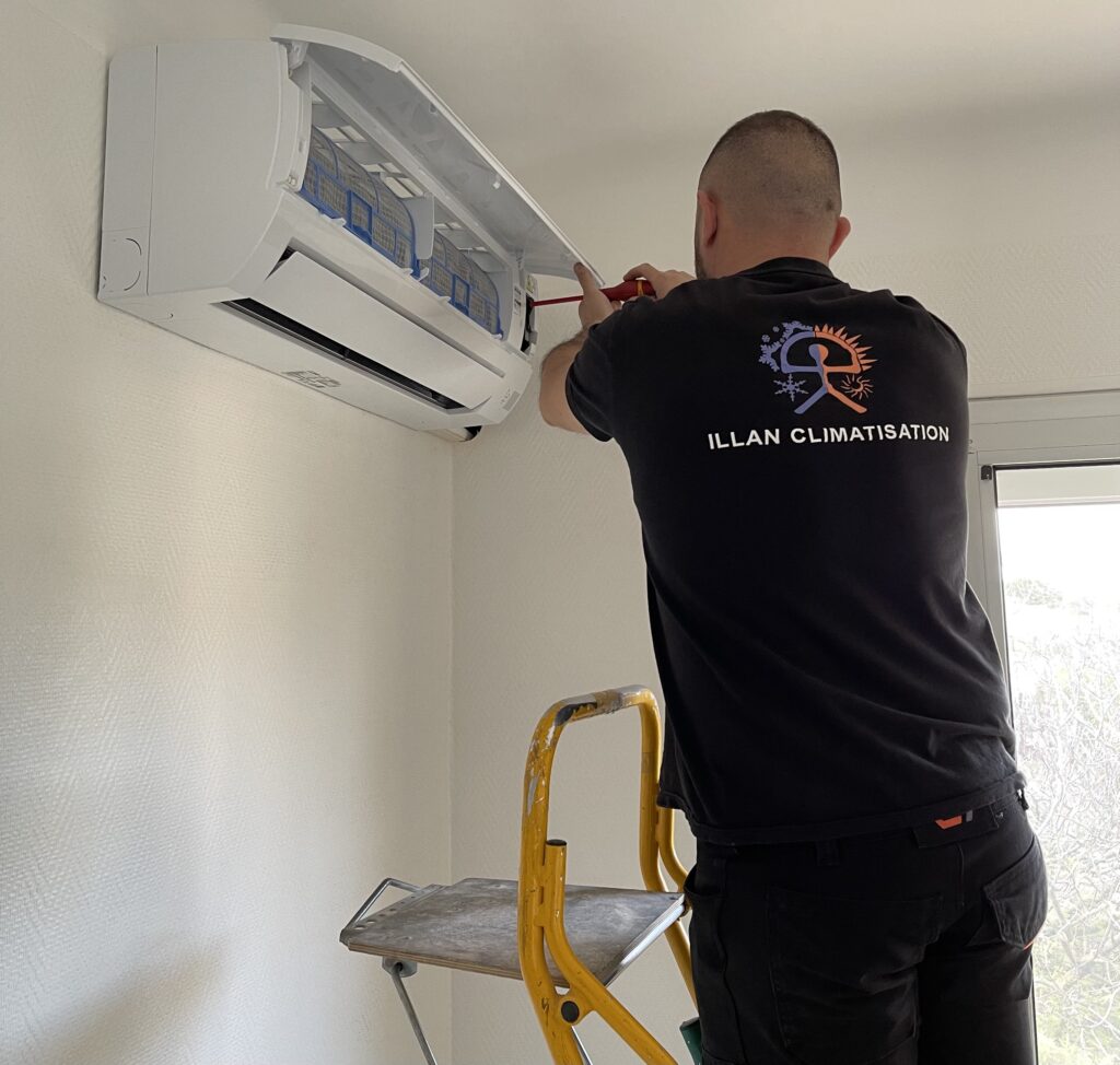 Maintenance Climatiseur : Illan Climatisation en Travail Technicien sur escabeau réparant un climatiseur mural intérieur, portant un t-shirt noir avec le logo et 'ILLAN CLIMATISATION'.