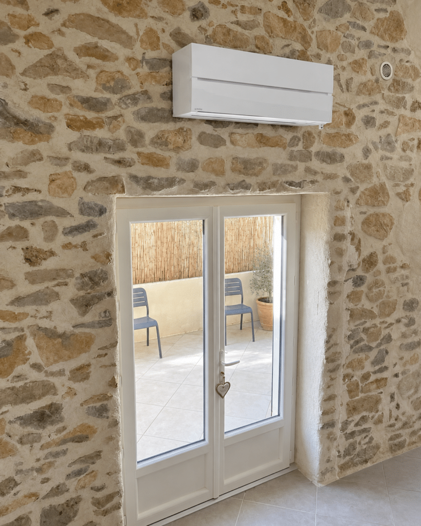 Installation de climatiseur sur mur en pierre apparente Climatiseur blanc sur mur en pierre apparente surplombant une porte-fenêtre ouvrant sur un patio.